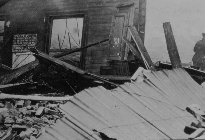 Black and white photograph from the aftermath of the Black Tom explosion of one wall standing surrounded by bricks and wooden debris